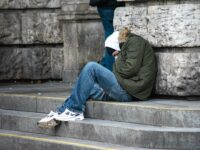 Ein obdachloser Mann sitzt auf einer steinernen Treppe, angelehnt an eine Mauer. Er trägt eine dicke Winterjacke, eine Mütze und Sportschuhe.