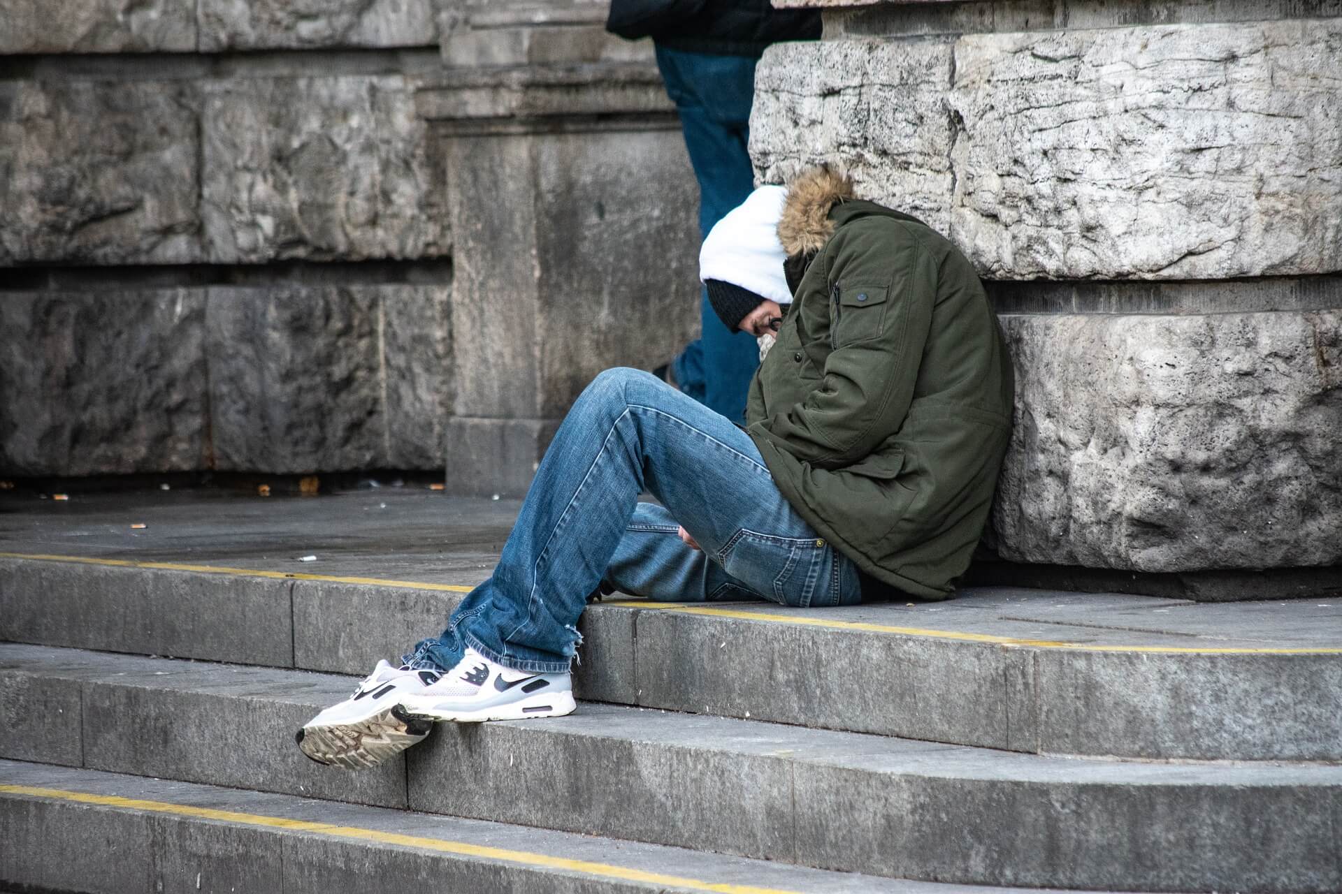 Ein obdachloser Mann sitzt auf einer steinernen Treppe, angelehnt an eine Mauer. Er trägt eine dicke Winterjacke, eine Mütze und Sportschuhe.